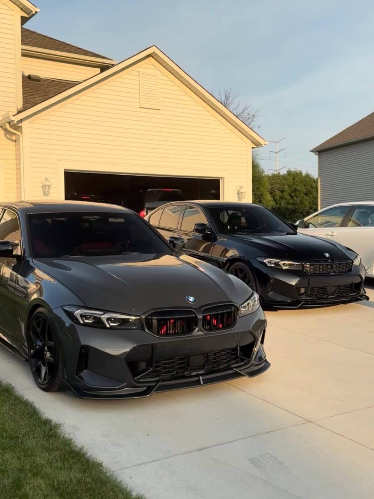 Two modified BMW sedans and a white sedan parked in a driveway in front of a house at golden hour.