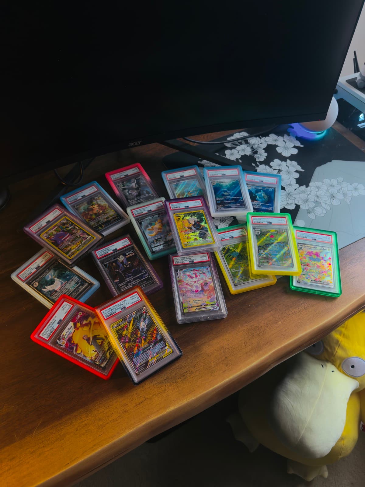 A fanned-out spread of graded Pokemon trading cards on a wooden desk, with a plush Psyduck toy in the corner.